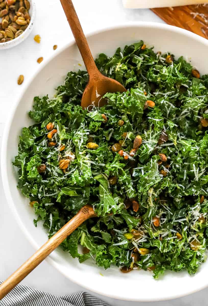 Mixed kale salad in a white salad bowl with wooden serving spoons in the bowl. 