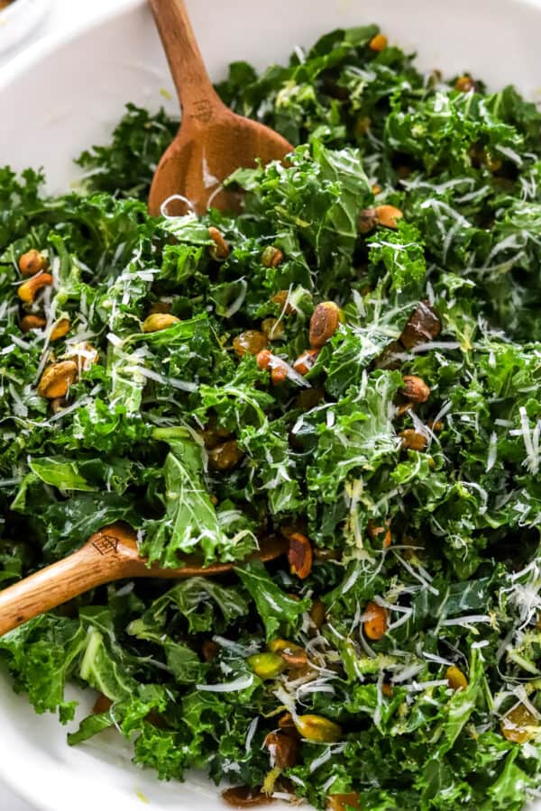 Close up of wood salad spoons mixing a kale salad in a white bowl.