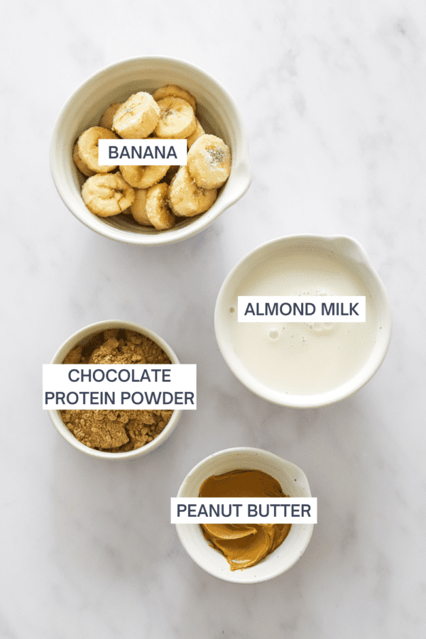 Frozen sliced banana in a bowl with a bowl of protein powder, almond milk, and bowl of peanut butter in front of it with labels over each ingredient.