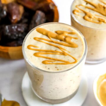 Two glasses filled with a white, creamy smoothie topped with peanut butter drizzle with a brown bowl of whole dates behind it.