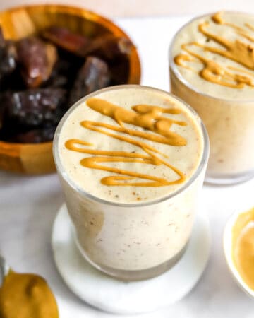Two glasses filled with a white, creamy smoothie topped with peanut butter drizzle with a brown bowl of whole dates behind it.