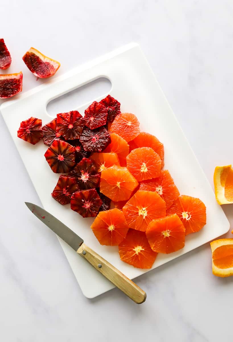 Sliced, peeled orange and red oranges on a white cutting board with a pairing knife on the board next to it.