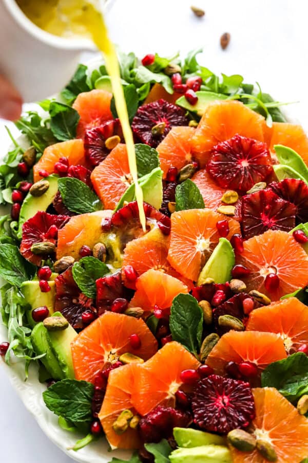 Hand pouring yellow dressing over a bed of greens topped with sliced oranges, avocado, nuts and pomegranate seeds.