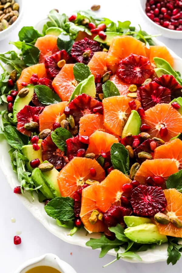 Platter full of a citrus salad with sliced avocado and pomegranate seeds on it and more behind it.