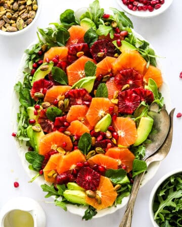 Citrus salad on a oval platter with some pistachios and pomegranate seeds behind it and some arugula in a bowl in front of it.