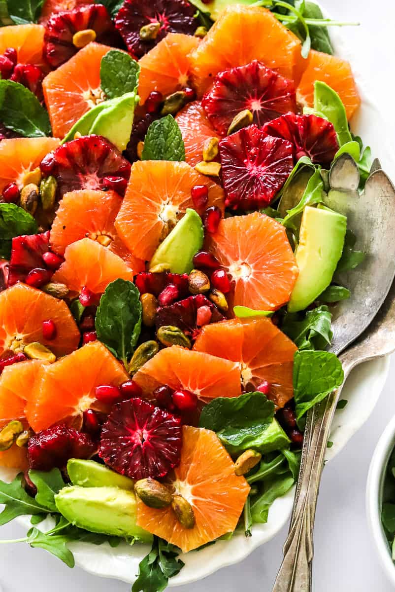 Arugula topped with sliced citrus, avocado, pomegranate seeds and pistachios with silver serving spoons on the platter.