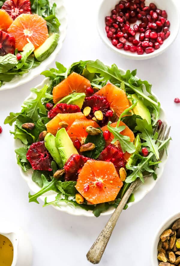Plate full of greens topped with sliced orange and red oranges, slice avocado and pomegranate seeds with more pomegranate seeds in a bowl behind it.