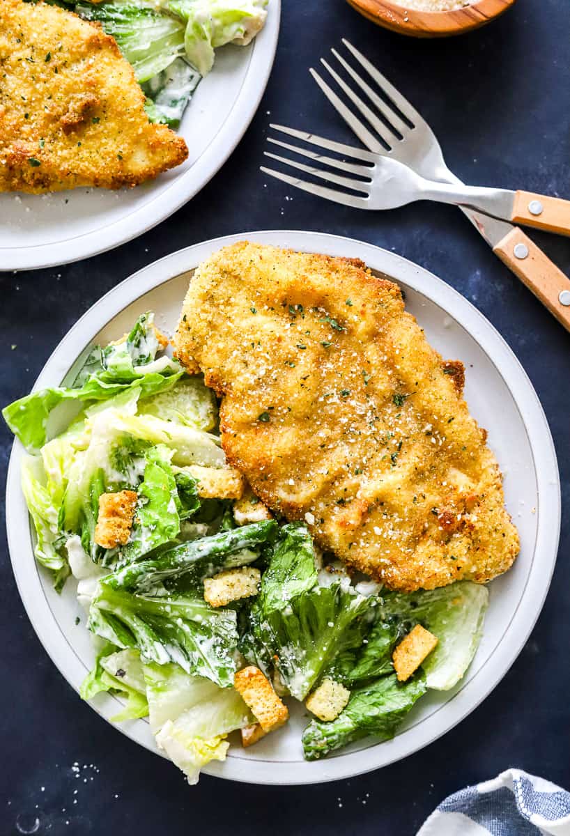 Air fryer chicken cutlet on a round grey plate with a green salad next to it and another plate of it behind it.