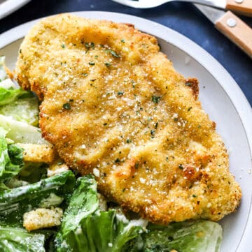 Golden breaded chicken on a plate with Caesar salad next to it and another plate and some forks behind it.