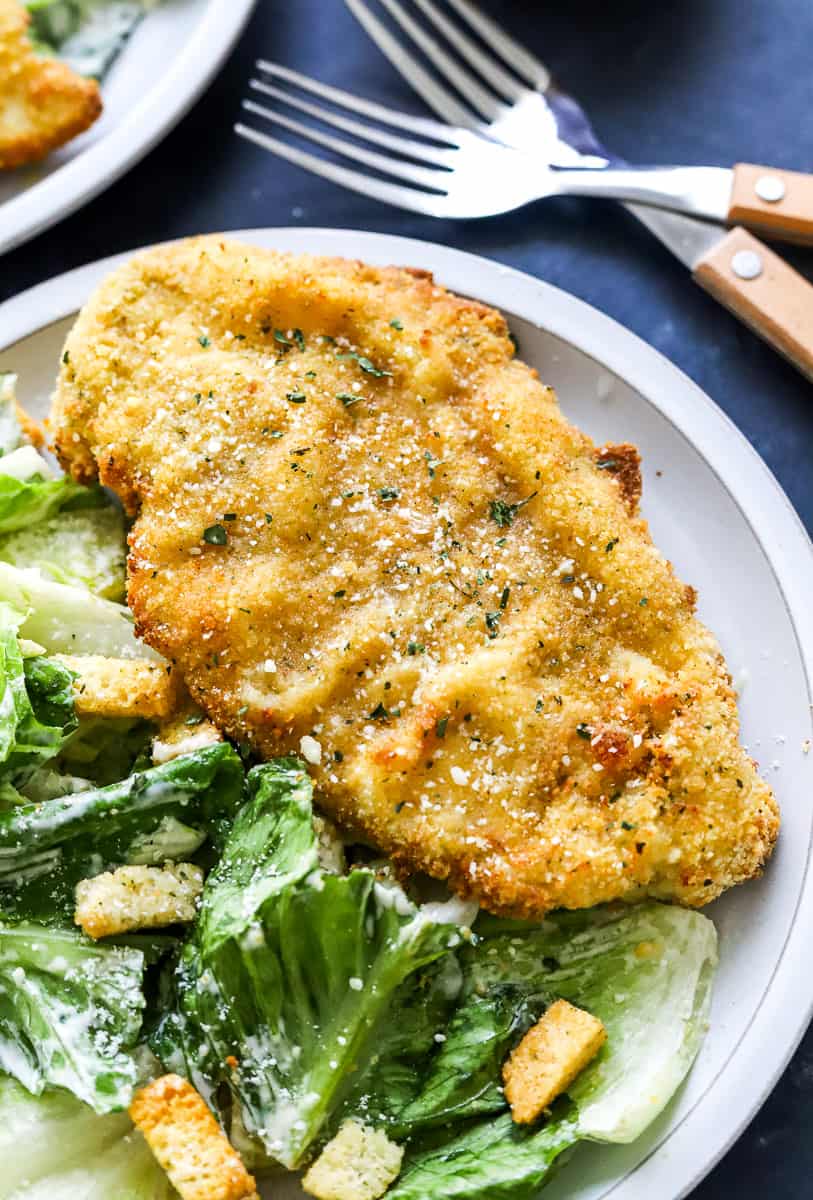 Golden breaded chicken on a plate with Caesar salad next to it and another plate and some forks behind it.