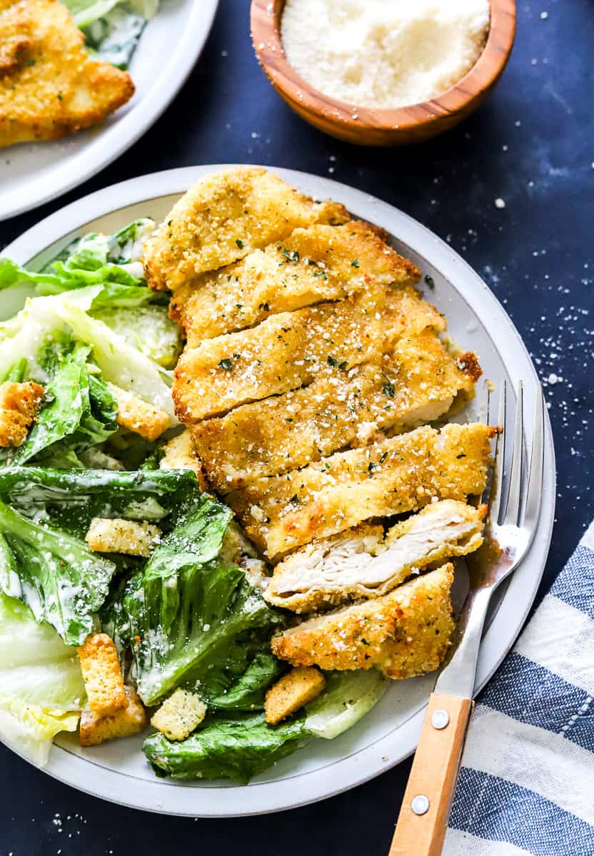Plate with a sliced pieces of crispy breaded chicken on it next to some Caesar salad with fork on the plate and a bowl of parmesan cheese and another plate behind it.