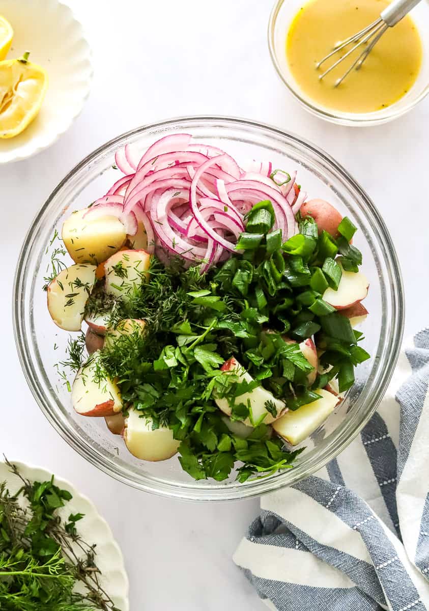 Cooked. chopped red potatoes in a bowl with green herbs, and sliced red onion with a bowl of mustard dressing and squeezed lemon behind it and a plate of herbs and a blue and white stripped towel in front of it.