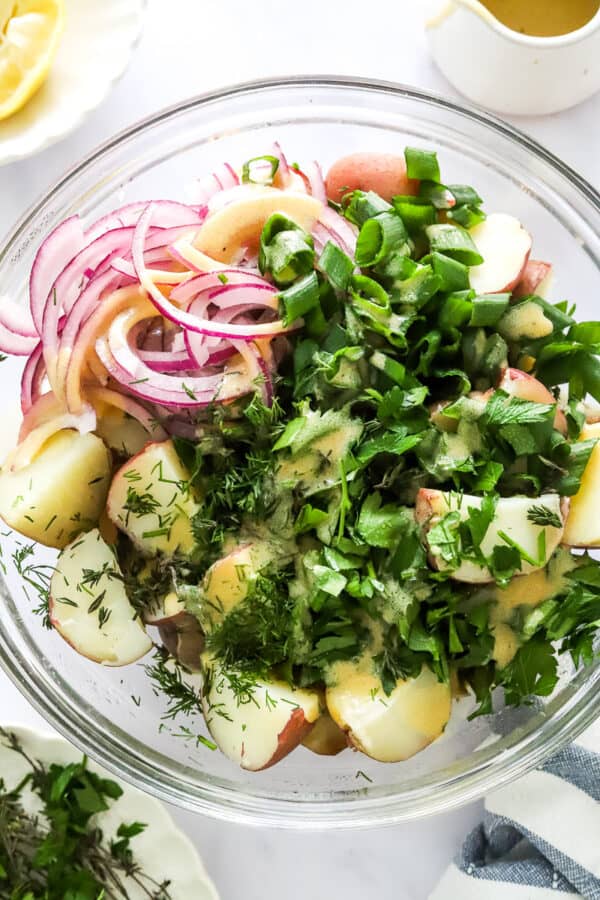 Glass bowl filled with cooked, chopped potatoes, herbs, sliced red onion and topped with a mustard dressing.