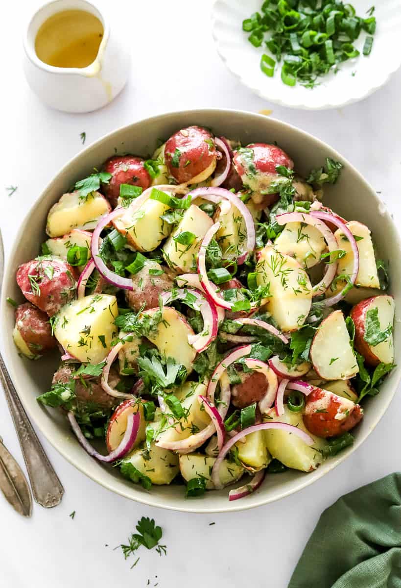 Bowl filled with healthy potato salad topped with sliced red onion and herbs with more herbs and mustard dressing behind it.