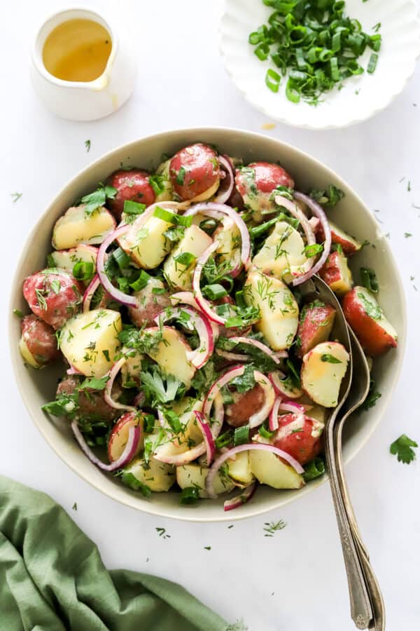 Red potato salad in a bowl topped with sliced purple onion and dill with serving spoons in the bowl with more dressing and chopped green onion behind it.