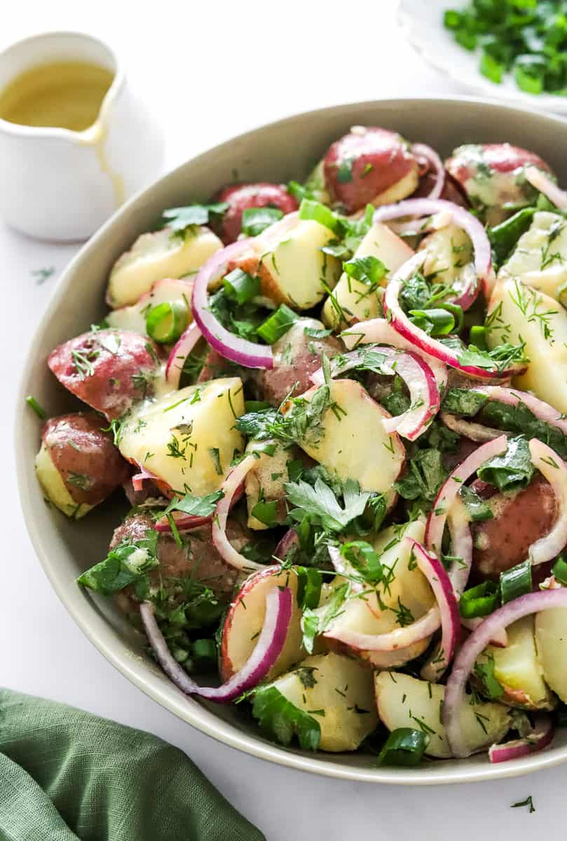 Quartered, cooked red potatoes mixed with a creamy mustard dressing, sliced red onion and fresh herbs in a bowl with more dressing and herbs behind it and a green towel in front of it.