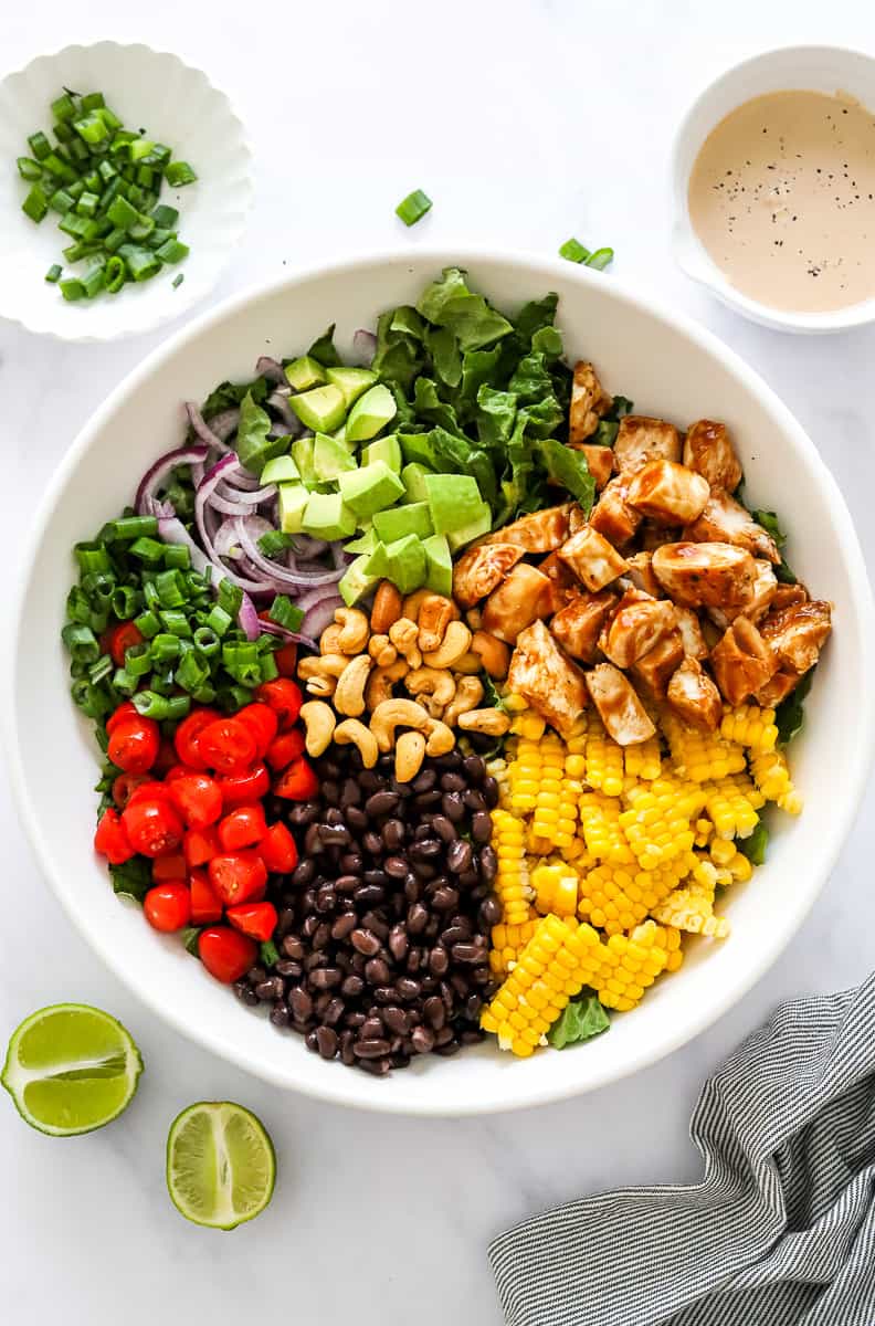 Large white bowl filled with chopped chicken, lettuce, avocado, red onion, green onion, tomatoes, corn and black beans. With a bowl of orange creamy dressing and a bowl of scallions behind it and a sliced lemon and striped towel in front of it.