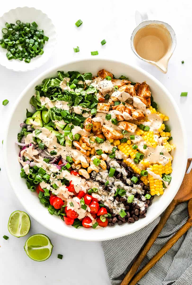 Large white bowl filled with chopped greens, veggies, and chicken topped with bbq dressing with a bowl of chopped scallions and more dressing behind it.