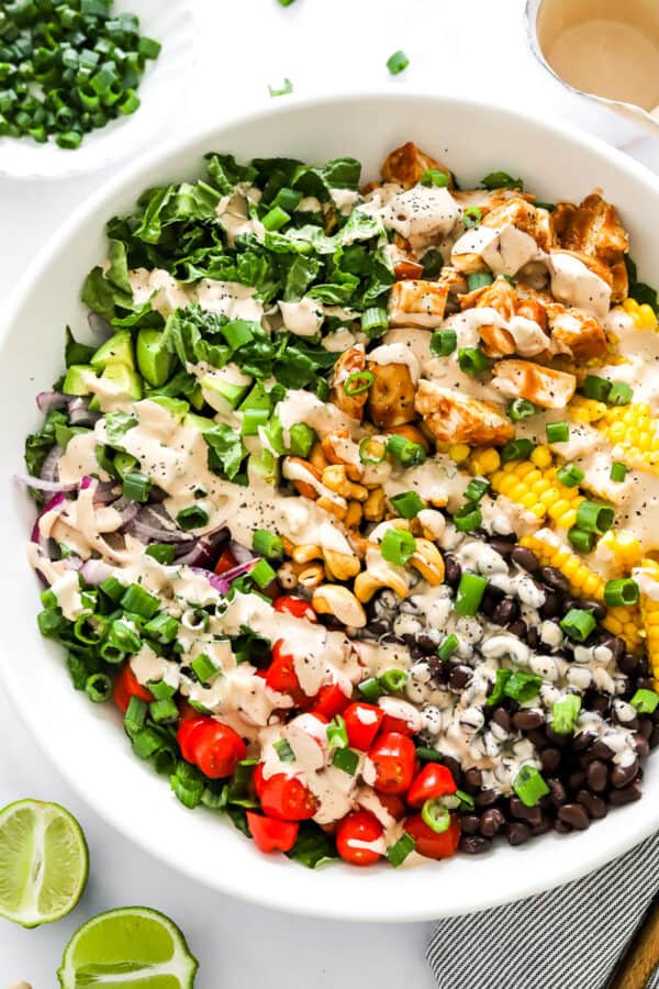 Large salad bowl filled with chopped lettuce, bbq chicken, corn, black beans, tomatoes and nuts with a drizzle of creamy bbq dressing over it with a bowl of chopped green onions and more dressing behind it and a sliced lime in front of it.