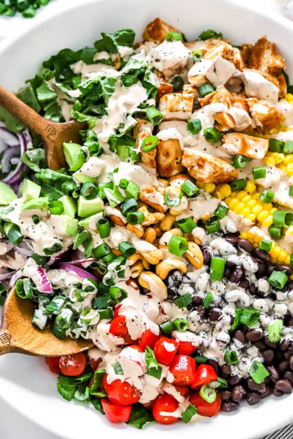 Close up of a creamy salad with chicken and fresh chopped veggies in a white, round salad bowl with wood serving spoons in it.