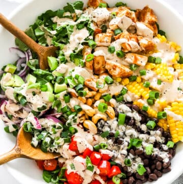 BQ Chicken in a bowl with some chopped greens, corn, black beans, tomatoes and avocado topped with a drizzle of a creamy salad dressing with wood serving spoons in the bowl.