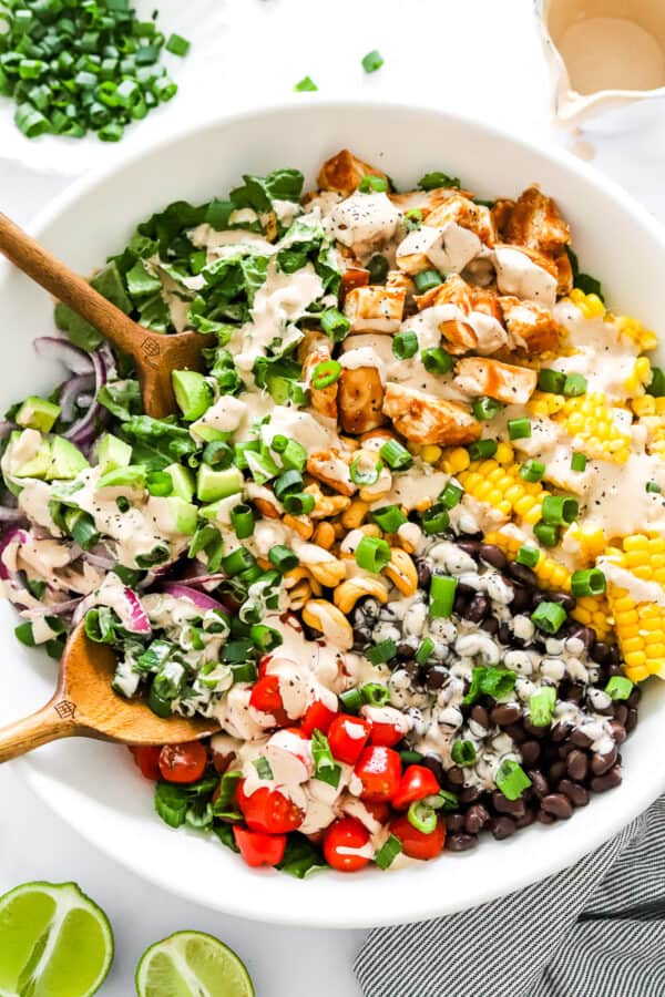 BQ Chicken in a bowl with some chopped greens, corn, black beans, tomatoes and avocado topped with a drizzle of a creamy salad dressing with wood serving spoons in the bowl.