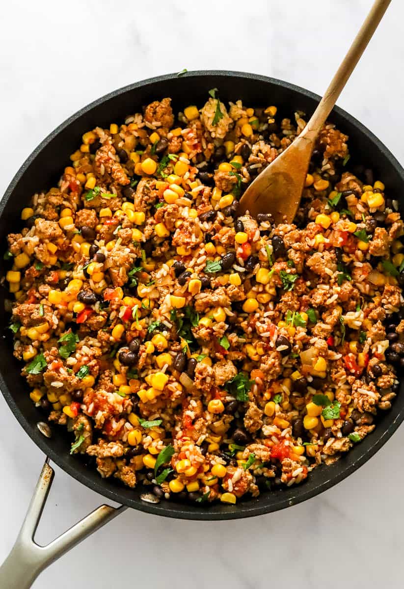 Cooked ground turkey, with corn, black beans and rice in a skillet with a wooden spoon in the pan.