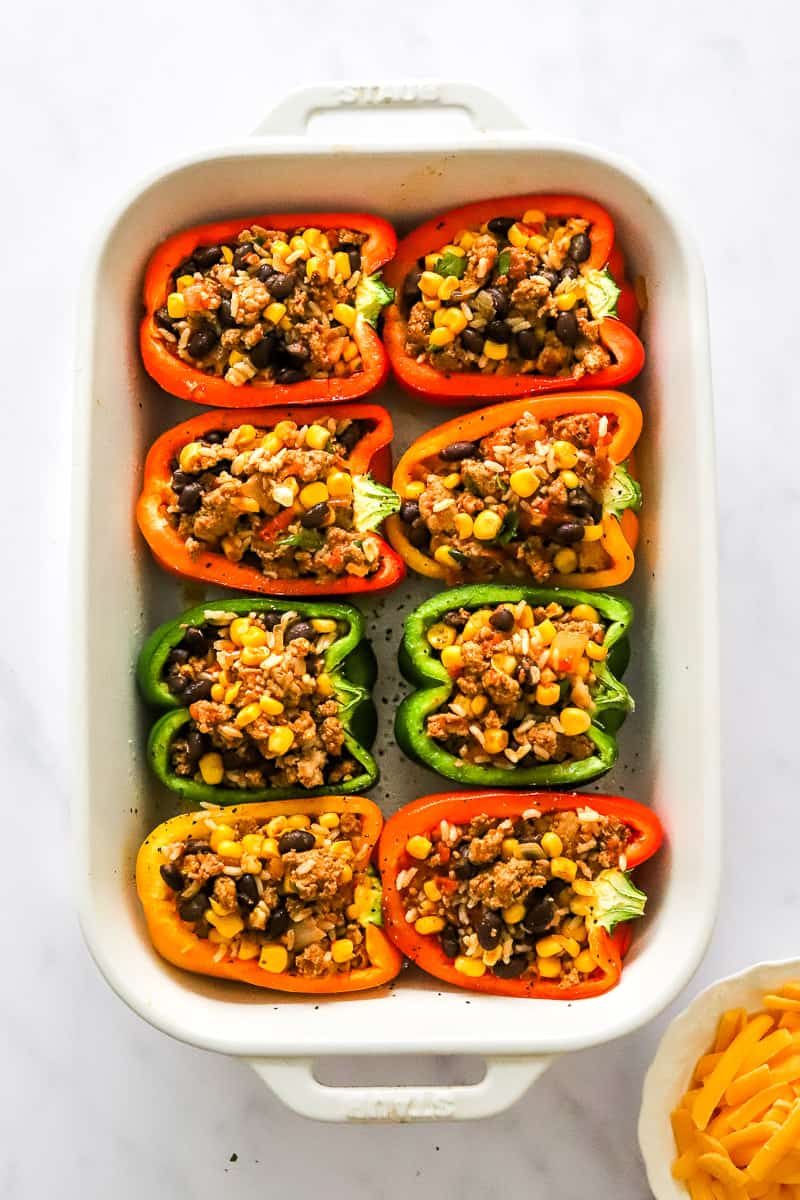 Multi colored bell peppers filled with a ground turkey veggie mixture in a white baking dish.