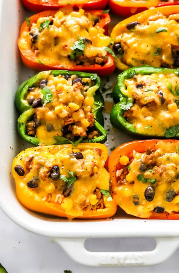 Multiple coloured bell pepper halves filled with a meat and corn mixture topped with melty cheese in a white pan.