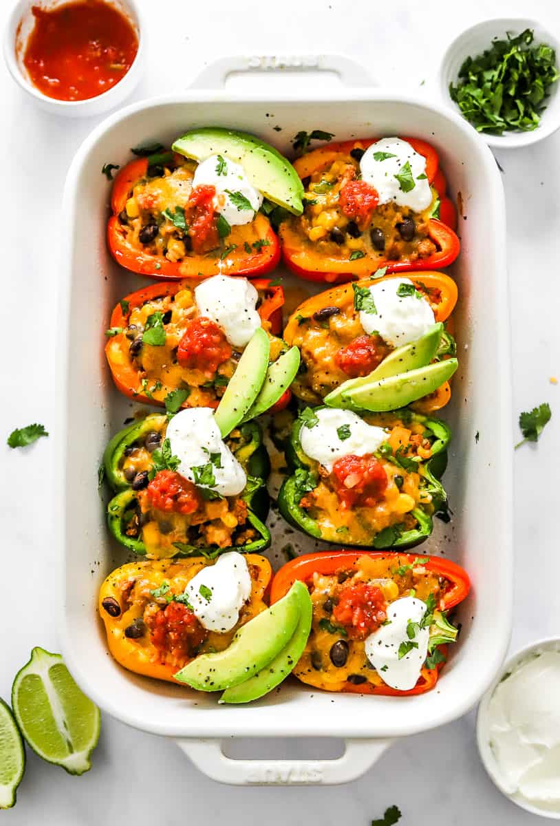 White baking dish filled with taco stuffed bell peppers topped with melted cheese, greek yogurt, salsa, and sliced avocado with more toppings in bowls around it.