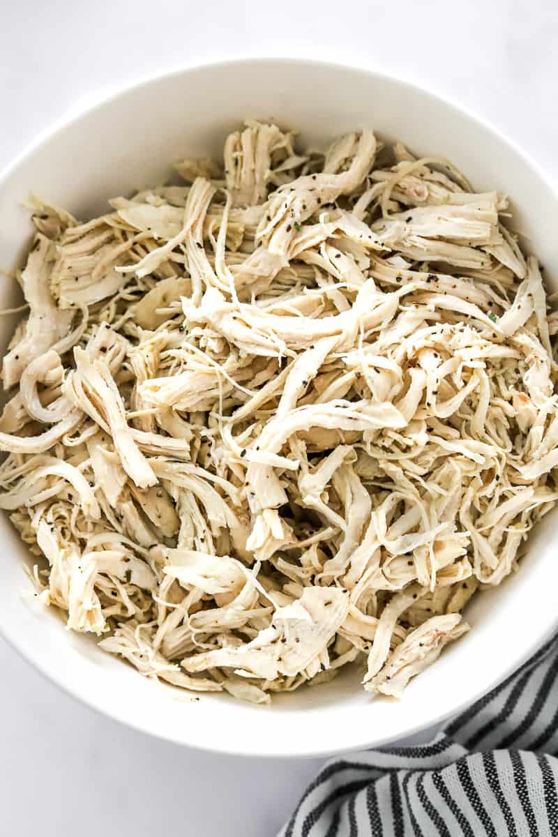 Serving bowl filled with cooked chicken that is shredded with a striped towel in front of it.