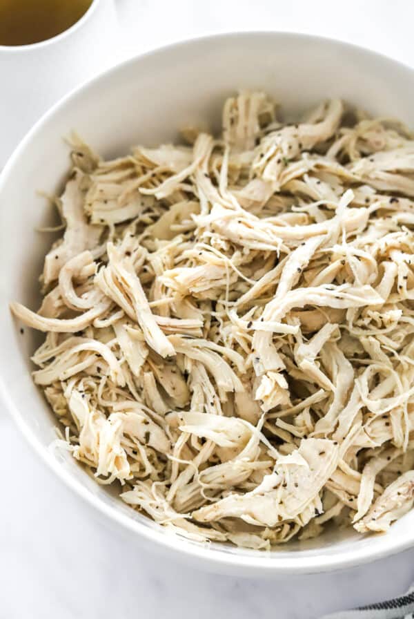 Instant pot shredded chicken breast in a white round bowl with some stock in a pitcher behind it.