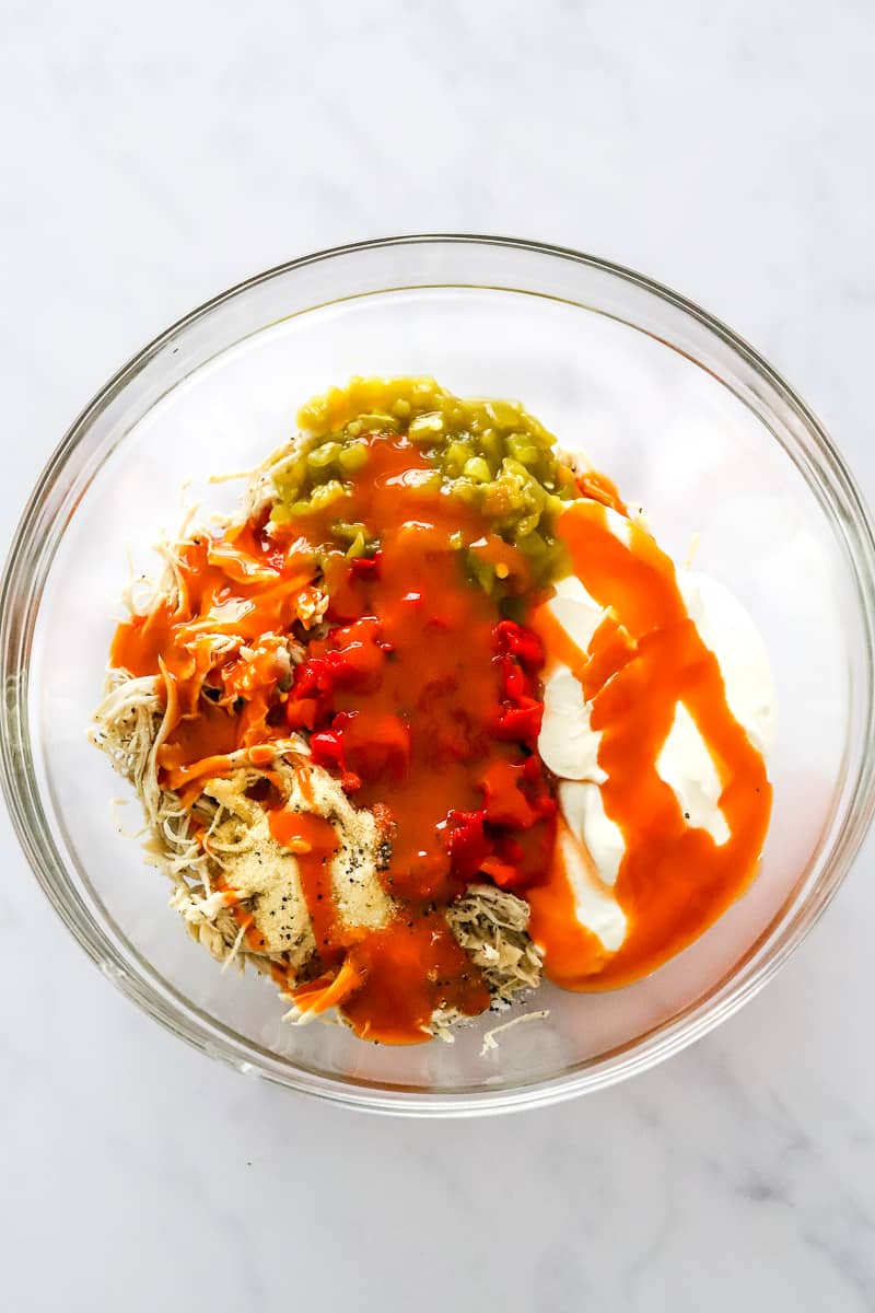 Glass mixing bowl filled with cooked shredded chicken, green chilis, red pepper, creamy cheese and cottage cheese drizzled with buffalo sauce.