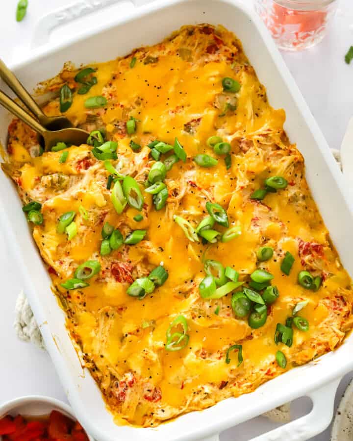 White dish with healthy buffalo chicken dip topped with melted cheese and sliced green onion on top of it with a jar of carrot sticks behind it and a bowl of red pepper in front of it.