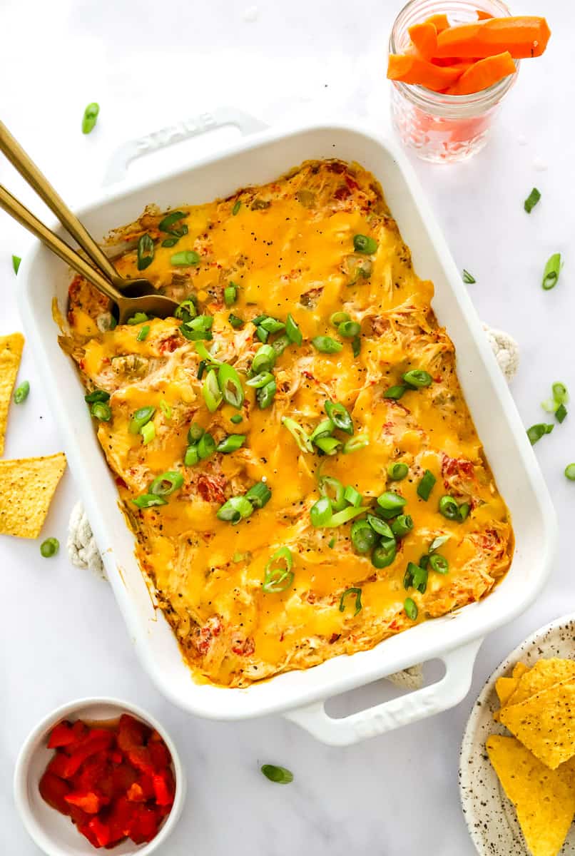 Creamy chicken dip topped with melted cheddar cheese and scallions in a white baking pan with serving spoons in the pan with a plate of tortilla chips and bowl of chopped peppers in front of it and glass jar of carrot sticks behind it.