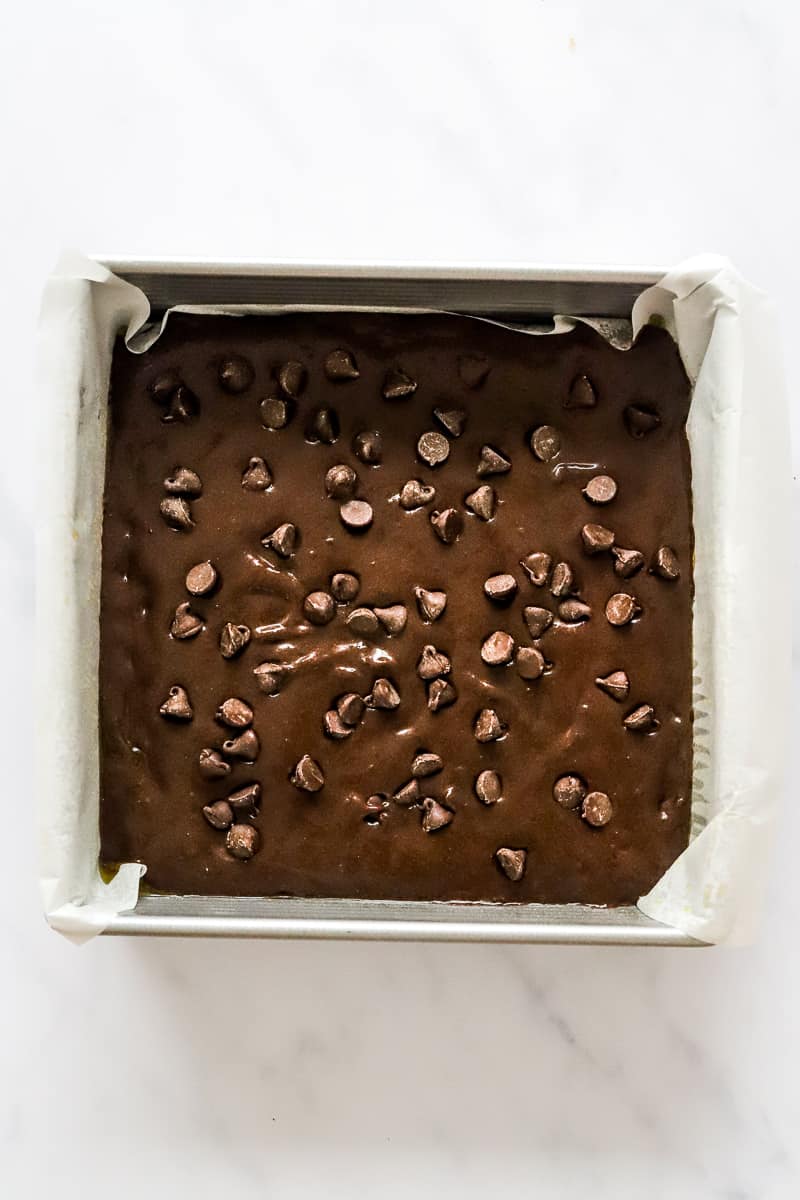 Uncooked brownie batter topped with chocolate chips in a metal baking dish that is lined with white parchment paper.