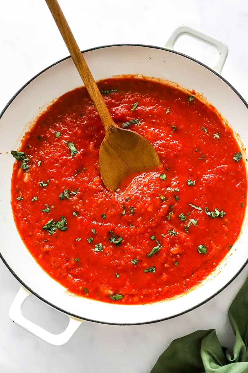 Pan of cooked tomato sauce with a wooden spoon in the sauce and basil on top of it with a green towel in front of it.