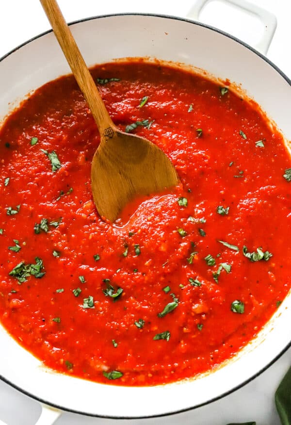 Cooked san Marzano tomato sauce in a white pan topped with basil with a wood spoon in the pan.