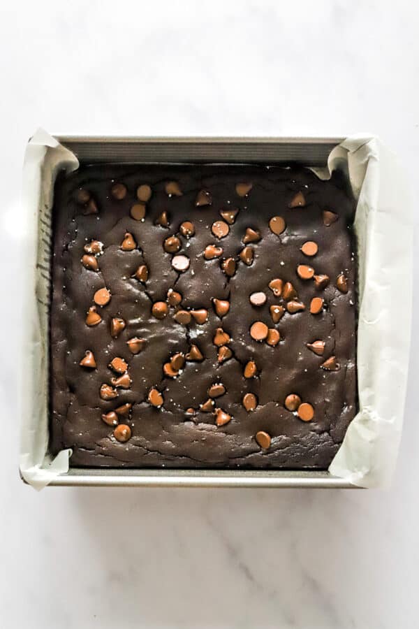 Square metal pan lined with white paper filled with baked avocado black bean brownies topped with chocolate chips.