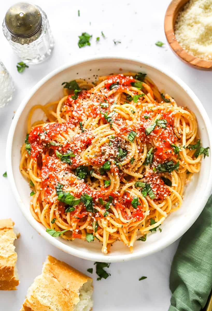 Noodle bowl filled with spaghetti tossed in tomato sauce with chopped basil on top of it with a piece of bread and a green towel in front of it and a bowl of grated parmesan and a salt shaker behind it.