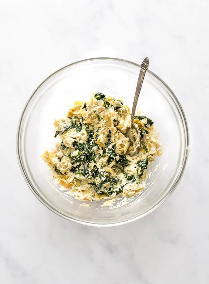 Artichoke, spinach and cheese filling in a round glass bowl with a silver spoon in the bowl.