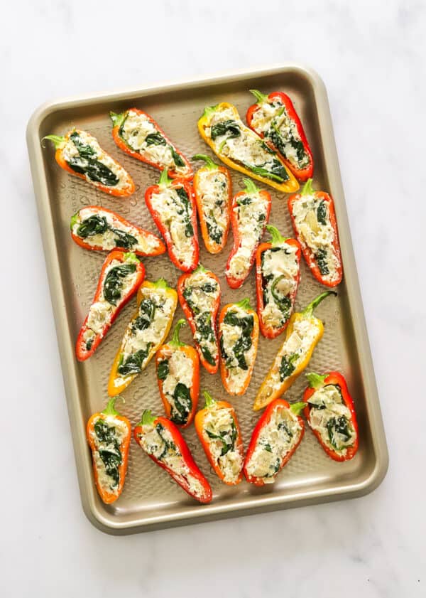 Baking tray filled with small colourful pepper halves that are filled with cheese and spinach filling.