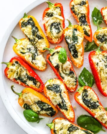 Round plate filled with a bunch of small sweet peppers filled with a cheese and spinach filling with basil leaves on the plate.