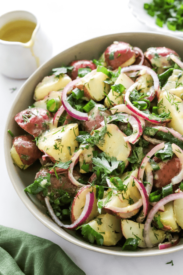 Bowl of herby healthy potato salad with more dijon dressing behind it and a green towel in front of it.