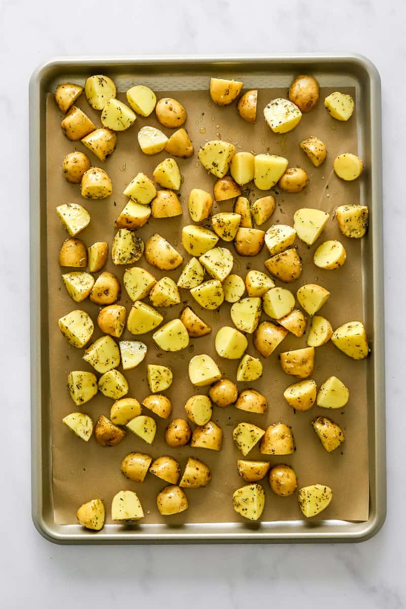 Baking sheet lined with brown parchment paper with raw seasoned cut baby yellow potatoes spread on it.