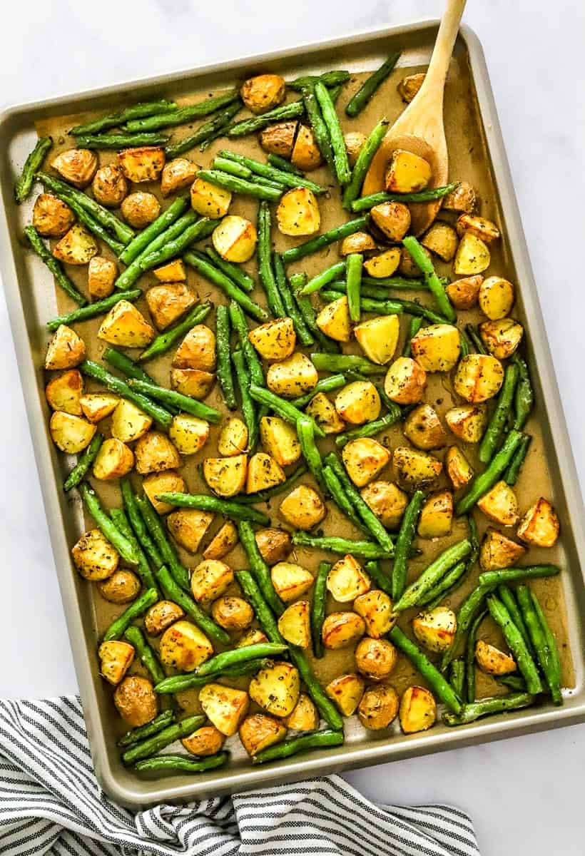 Large baking pan filled with roasted baby potatoes and green beans with a wooden spoon on the baking sheet and a striped towel in front of it.