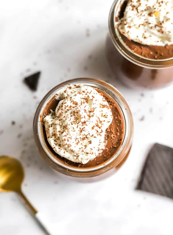 Two glass jars of chocolate cream topped with whipped cream with a spoon and a bar of chocolate in front of them.