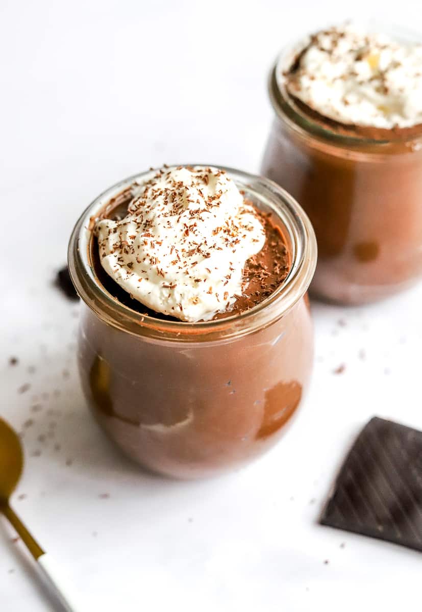 Glass jar filled with chocolate mousse with whipped cream and shaved chocolate on the top of it with another jar of it behind it and a spoon in front of it.