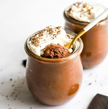 Jar filled with vegan chocolate mousse topped with whipped cream with a spoon in the jar and another jar of the mousse behind it with a square of chocolate in front of it.