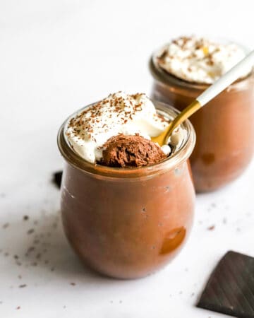 Jar filled with vegan chocolate mousse topped with whipped cream with a spoon in the jar and another jar of the mousse behind it with a square of chocolate in front of it.
